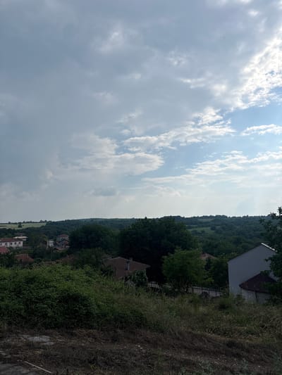This image, presented rotated 90 degrees clockwise, displays a panoramic daytime landscape, likely set in Kralevo, Bulgaria. The upper portion of the frame is dominated by a vast sky, filled with a dynamic mix of white and grey cumulus clouds, with scattered patches of clear blue sky peeking through.
In the middle ground, a long, undulating line of densely forested hills or mountains stretches across the horizon, appearing dark green due to the distance and heavy tree cover. Below this natural boundary, areas of lighter green suggest open fields or meadows.
The lower and right-hand side of the scene features several houses with distinct roof colors, including reddish-brown and grey, integrated into the natural environment. These dwellings are nestled among lush green trees and thick bushes. The immediate foreground on the right exhibits dry, overgrown grass and scrub, interspersed with areas of bare earth, indicating possibly uncultivated land.
The lighting suggests it is daytime, under a partially cloudy sky. No people, animals, or specific activities are discernible in the tranquil rural setting. There is no visible text in the image.