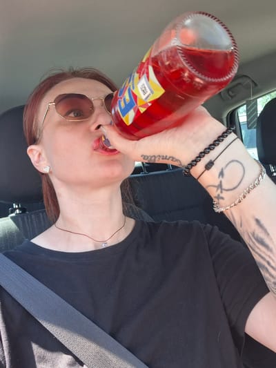 A woman with red hair and sunglasses is in the driver's seat of a car, drinking from a red bottle. She is wearing a black t-shirt and a silver heart-shaped earring. There are tattoos on her left arm. A seatbelt is visible across her chest. The background shows car interior and a glimpse of the outside, suggesting it is daytime. There is a hanging air freshener visible in the car.