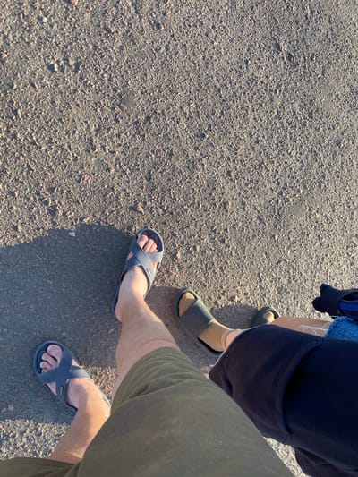 The image shows a ground-level perspective of two pairs of legs standing on a dusty, asphalt-like surface. The surface appears to be a path or road, with small stones and debris scattered across it. Sunlight casts a distinct shadow on the left side of the frame.

The person on the left is wearing dark grey slide sandals and olive green shorts. Their legs are bare from the knee down. The person on the right is wearing dark grey slide sandals and black shorts. They are also wearing light tan socks and a dark blue item of clothing that appears to be a jacket or hoodie. Part of their legs are also visible.

The scene is likely outdoors, given the natural lighting and the texture of the ground. The location in Yablunivka, Ukraine is not visually discernible from the image itself, but the description focuses on the elements present.