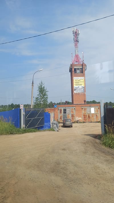 The image depicts a rural scene in Khokhryaki, Russia, dominated by a tall brick tower topped with a telecommunications mast. The tower has several antennas and is red and white. A yellow advertisement banner is attached to the side of the tower. At the base of the tower, a row of brick garages with rusty doors is visible. A dark-colored car is parked in front of one of the garages. To the left of the garages, there is a blue metal fence and a gate with a circular sign indicating a speed limit of 10. Bags of what appear to be construction materials are piled near the fence. A street lamp stands to the left of the fence. The ground is a dirt road. The sky is blue with some wispy clouds. Trees and foliage surround the area, suggesting a location on the outskirts of a settlement.