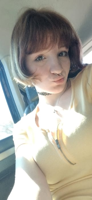 A single individual, identified as a female, is depicted in a close-up, front-facing perspective, likely a selfie. She has reddish-brown, chin-length hair with lighter blonde or light brown tips, styled with bangs across her forehead. Her eyes are light-colored, and she has visible freckles on her face. A silver-toned septum piercing is visible in her nose, and her lips are puckered. She wears a black choker necklace and a silver-colored chain necklace with a small, dark pendant. Her attire consists of a light yellow short-sleeved top with a metallic zipper running vertically down the front, partially unzipped.
The setting appears to be the interior of a vehicle, possibly in the front passenger seat. A dark, upholstered seat or seatback is visible behind her left shoulder. To the left of the frame, a window frame and bright outdoor light are apparent, causing significant overexposure in that area and casting strong illumination on the subject's left side. The upper part of the background, above her head, is a light, indistinct surface, possibly the vehicle's ceiling. The image provides no specific visual cues to confirm the stated location of Bistrice, Montenegro.