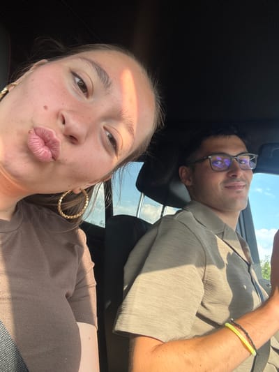 The image shows a selfie taken inside a vehicle, likely a car. Two individuals are visible, a woman in the foreground and a man in the background. The woman, positioned on the left, leans in towards the camera with her face angled to the side, puckering her lips. She has fair skin, brown eyes, and is wearing a brown t-shirt and large gold hoop earrings. Sunlight casts a warm glow on her face. The man, on the right, is wearing glasses and a light-colored collared shirt. He is looking towards the camera with a slight smile. His arm is visible, and he is wearing a dark bracelet with a neon yellow element. The interior of the car is dimly lit, with the dashboard and seats suggesting a typical vehicle interior. The background outside the car window shows a clear blue sky with some greenery. The scene appears to be a casual moment captured while traveling. The mention of Rupkite, Bulgaria, in the prompt does not correlate with any visual information present in the image.