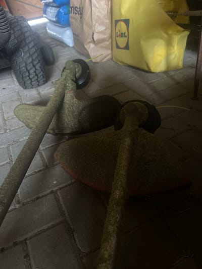 The image depicts an outdoor scene, likely a storage area or garage, paved with interlocking bricks. Two grass trimmers, heavily covered in grass clippings, are the primary focus. In the background, to the left, a portion of a large tractor tire is visible, along with stacked blue and grey plastic-wrapped items and a brown paper bag with the "Mumsay" logo. To the right, a bright yellow reusable shopping bag with the Lidl logo is prominent. The lighting is dim, suggesting it might be evening or an indoor area with minimal light. The overall impression is one of a yard or garden equipment storage area, possibly in Dubingiai, Lithuania, given the mention of the city.