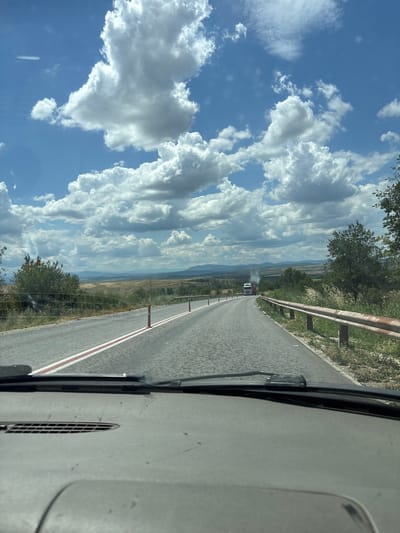 The image captures a view from inside a vehicle traveling on a paved road in Radomirtsi, Bulgaria. The road has a textured surface and is marked with a continuous white line with a red stripe along its edge. Orange traffic cones are visible along the right side of the road. On the left, there is a metal guardrail with visible rust, and beyond it, vegetation and trees. In the distance, rolling hills and mountains are visible under a bright blue sky with numerous fluffy white clouds. A white truck with a red trailer is seen in the distance on the road, emitting a plume of smoke. The foreground shows the interior of the vehicle, including the dashboard and windshield wipers. The overall environment appears to be rural and open.