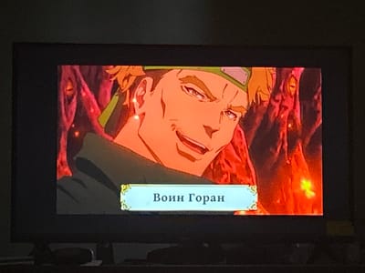 The image shows a close-up of a television screen displaying an anime-style character with orange hair and a green headband. The character has a smug expression and is looking to the right. Below the character is a light blue banner with decorative gold accents on the left and right. Inside the banner, in dark blue Cyrillic text, are the words "Воин Горан" which translates to "Warrior Goran". The background of the image on the screen is fiery red and orange, with indistinct shapes that suggest a dangerous or chaotic environment. The television screen is black and appears to be in a dimly lit room, with a subtle reflection visible on the screen's surface. There is no indication of Florești, Moldova in the image.