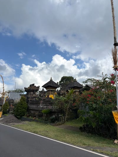 The image shows a traditional Balinese temple with intricate stone carvings and thatched roofs, located in Susut, Indonesia. The temple is situated along a road, with lush greenery and vibrant red flowers surrounding it. The sky is partly cloudy, with a mix of blue sky and white clouds. A tall, decorative bamboo pole with woven decorations stands on the right side of the frame. The road in the foreground is paved and has a white dividing line. The scene depicts a typical rural landscape in Bali, showcasing the island's rich cultural heritage and natural beauty.