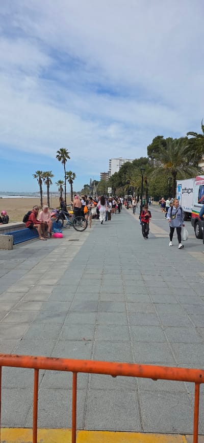 The image depicts a street scene in Benicàssim, Spain, with a view of the coast on a day with a partially cloudy sky. Palm trees line the street alongside buildings in the background. A group of people appears to be walking or gathering on the sidewalk. A few people are seated on benches near the beach. A bicycle is parked near the sidewalk. An orange barrier is visible in the foreground. An ambulance is parked on the side of the road further ahead. The sidewalk is paved with gray tiles.