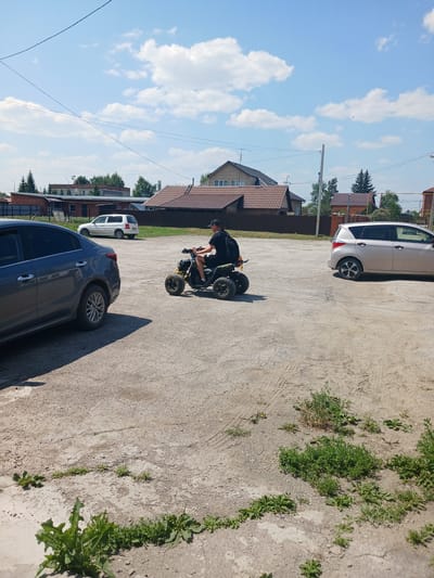 In Prokudskoye, Russia, on a sunlit day under a partially cloudy sky, a young person wearing a black t-shirt and a black baseball cap rides a yellow and black ATV. The ATV is positioned in the center-right of the image, on a light-colored, gravelly asphalt surface. The rider appears to be moving forward. To the left of the ATV, a dark gray sedan is parked, its rear visible. Further back, a white SUV is parked. The background features residential buildings with tiled roofs, some trees, and utility poles. Green vegetation, including weeds and grass, is scattered across the foreground and parts of the parking area. The overall scene depicts a casual outdoor setting.