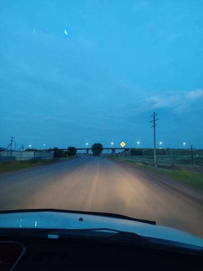 This media file shows a first-person perspective shot from inside a vehicle driving on a road at dusk. The sky is a deep blue with a few wispy clouds, indicating the transition from daylight to night. The headlights of the car illuminate the road ahead, creating streaks of light that suggest motion.
The scene is rural, with grassy fields and scattered trees visible on either side of the road. In the distance, a highway overpass with streetlights can be seen, along with power lines and what appears to be an industrial or agricultural facility on the left. A diamond-shaped road sign is also visible on the overpass, illuminated by streetlights. The overall atmosphere is quiet and serene, typical of a twilight drive in a less populated area. The location is identified as Derköl, Kazakhstan. There is no visible text within the image.
