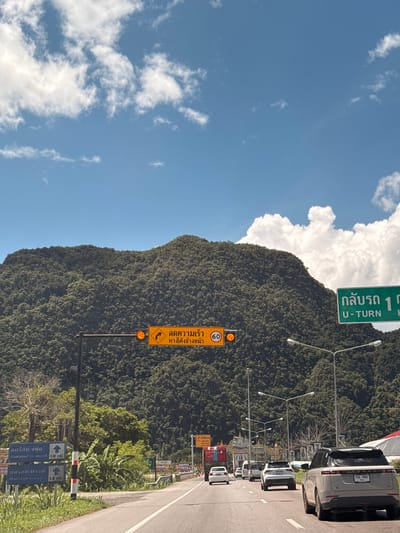 The image captures a scenic view of a mountain covered in lush green vegetation under a bright blue sky with scattered white clouds. A road runs through the foreground, with several cars driving on it. Traffic signs are visible, including one in Thai indicating "Reduce Speed, Elephant Crossing Ahead," with a flashing orange light. Another green sign indicates "U-TURN 1 km." The overall impression is of a natural landscape encountered on a journey through Phang-nga, Thailand, likely on a sunny day.