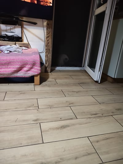 The image depicts an interior scene, likely a room, with a focus on the floor and a portion of a bed. The floor is covered in light-colored wood-look tile arranged in a staggered pattern. To the left, a portion of a bed is visible, covered with a pink and white crocheted blanket. A white t-shirt or similar garment is draped over the blanket. Behind the bed, a white wall is partially visible, with a wooden beam frame. A black television is mounted above this area, with cables running down the wall.
In the center of the image, a doorway or window area is visible. The glass is dark and partially obscured, but a faint, blurry outline of a light-colored animal, possibly a dog, can be seen just inside. The doorway or window frame is white.
The overall impression is that of a domestic interior, possibly a bedroom or living space. The specific location in Ungheni, Moldova, cannot be determined from the visual information provided in the image.