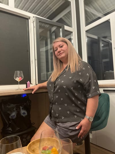 The image depicts an indoor scene in Kamyennoye, Russia, featuring a single adult female in what appears to be a residential or private bar-like setting.
**Subject:**
A female individual, estimated to be between 35-55 years old, with light skin and shoulder-length blonde hair, is seated on a high stool. She is wearing a dark grey short-sleeved collared shirt patterned with numerous small white diamond shapes and dark grey shorts. A silver-colored watch with a round, light-colored face and a mesh band is visible on her left wrist. Her fingernails are manicured with a light polish. Her right hand rests on a wooden counter surface. She is looking directly towards the camera with a neutral expression.
**Environment and Objects:**
The background features multiple white-framed windows. The leftmost window panel has a dark blind or screen drawn. Other window panels show reflections of the interior and an overhead white slatted structure, possibly an awning or ceiling. A dark vertical beam separates some window sections.
On the wooden counter, to the left of the woman's hand, is a clear stemmed wine glass containing several translucent, multicolored cube-shaped objects (likely ice or decorative items) and a small, opaque pink object. Adjacent to the glass is a rectangular, pink and black object resembling a disposable vape pen.
Under the counter, to the left, a black plastic bag with a printed image of stylized animal eyes (blue irises) is visible, resting on a dark stand or small stool. A subtle blue glow emanates from beneath the counter.
In the foreground, on a lower wooden surface, two empty clear glasses and a wooden bowl filled with various colorful fruits (including grapes, berries, and orange segments) are present. A dark green upholstered high-backed stool or chair is partially visible to the right of the woman. The flooring appears dark. The overall lighting is artificial, suggesting evening or nighttime.