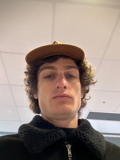 A fair-skinned male individual with curly brown hair and blue eyes is depicted in a close-up, low-angle frontal shot, looking directly at the camera with a neutral expression. He wears a brown baseball cap, positioned backward and slightly elevated, and a dark-colored jacket featuring a prominent, textured black collar with a visible metal zipper pull. The background consists of a white drop ceiling with distinct grid lines, typical of an indoor commercial or public building. A rectangular ventilation grille is partially visible in the upper right quadrant of the image. The specific location within Joinville-le-Pont, France, cannot be determined from the visual evidence presented.