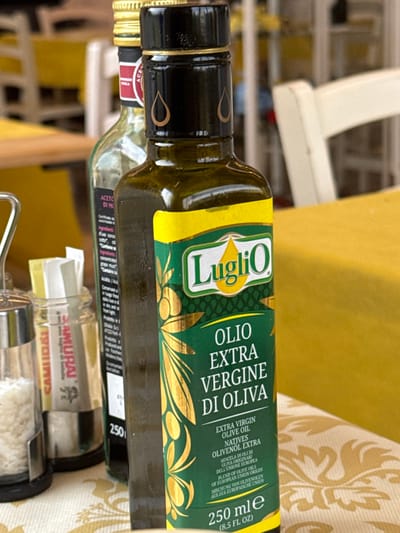 A close-up, eye-level photograph captures a dining table setup, presumably in a restaurant or trattoria in Anzola dell'Emilia, Italy. The primary subject is a dark green glass bottle of "LugliO Olio Extra Vergine di Oliva" (Extra Virgin Olive Oil), prominently labeled in green and yellow with gold olive branch motifs. The bottle features a black cap adorned with two golden teardrop shapes and indicates a volume of "250 ml (8.5 FL OZ)". Partially visible behind it is a taller, clear glass bottle with a gold cap, displaying a dark label with text "ACETO DI MO..." (likely referencing balsamic vinegar). To the left of the olive oil, a metal and glass shaker containing coarse white salt stands next to a small clear glass holder containing white packets or napkins, one of which exhibits red text, including "SAMURAI." The table is covered by a yellow tablecloth featuring a subtle, light-colored repeating pattern. Blurred in the background, out-of-focus yellow and white chairs and a wooden table edge indicate a lively, furnished dining environment. No individuals are present in the frame, and no actions or interactions are depicted.