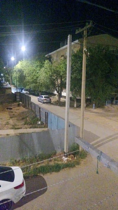 The image depicts a nighttime street scene in Maqat, Kazakhstan, with a focus on a residential area and its surrounding infrastructure. A street light illuminates the scene, casting a glow on a road where a silver sedan is parked. Adjacent to the road, a corrugated metal fence runs along a dirt area with sparse vegetation. Further in the background, a multi-story building with numerous windows is visible, partially obscured by dense green trees. Two concrete utility poles stand prominently in the foreground and midground, supporting overhead wires. Another white car is partially visible in the lower left corner of the frame. The overall impression is one of a quiet, functional urban environment at night.