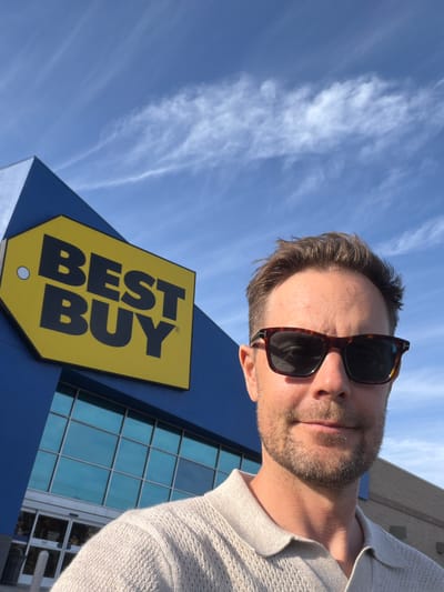 A medium-shot, low-angle selfie shows a man in his 30s or 40s standing in front of a Best Buy store. The man is wearing tortoiseshell sunglasses and a beige, cable-knit polo shirt. He has short, brown hair and light stubble on his chin and cheeks. The Best Buy store has a blue facade and a large yellow sign with black lettering that reads "BEST BUY". The store entrance, with glass doors and a blue sky visible through the windows, is in the background. The sky is bright blue with wispy white clouds. The location is identified as Westminster, United States of America, likely on a clear, sunny day.