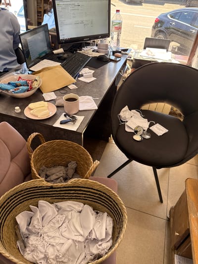 A wide shot captures an indoor scene, likely a small shop or office, with a window to the right revealing a street scene and parked vehicles. The foreground is dominated by two woven baskets. The larger basket, closer to the viewer, is filled with numerous small, white drawstring pouches. The smaller basket, positioned behind the first, contains a pile of grey fabric items. To the left, a portion of a pink upholstered chair is visible.

The mid-ground features a dark, modern office chair with a round seat and four legs. On the seat of this chair, several small white pouches, a compact mirror, and some rectangular cards are scattered. A dark grey desk runs horizontally across the image. On the desk are two computer monitors, a laptop, a keyboard, and a mouse. To the left of the monitors, a bowl filled with snacks and a bottle of water are present. A pink plate with sliced cheese and a mug of coffee sit on the desk, along with a set of car keys on a white napkin. An open brown envelope and various papers are also on the desk. The floor appears to be tiled.

The background, visible through the window, shows a paved street with a black car parked at an angle. Buildings are faintly visible further in the distance. In the upper left corner of the image, a person wearing a light blue shirt is partially visible, facing away from the camera and towards the desk area. Another person, wearing a lighter blue shirt, is visible in the upper left background, further away. The overall impression is of a workspace that is also preparing or distributing items, possibly related to a retail or service business. The location is identified as Sunzha, Russia.