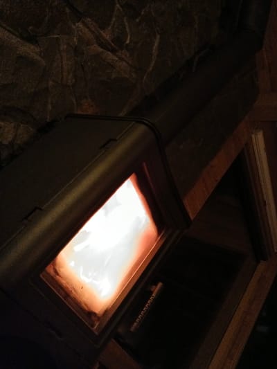 An indoor scene in Korostiv, Ukraine, depicts a dark-colored metal wood-burning stove or fireplace insert positioned at an upward diagonal angle within a dimly lit room. A bright, white and orange fire is actively burning within the stove's glass-fronted combustion chamber, serving as the primary light source. Above the stove, a wall constructed from dark, irregularly textured stone or rock is visible. A dark metal flue pipe extends diagonally upwards and to the right from the top of the stove. To the right of the stove and flue, dark wooden structural beams or framing are present, with a lighter vertical element visible in the far right, possibly part of a door frame or fixture. A dark, recessed area below the firebox shows a small, light-colored, ridged object partially obscured. No individuals are present in the image.