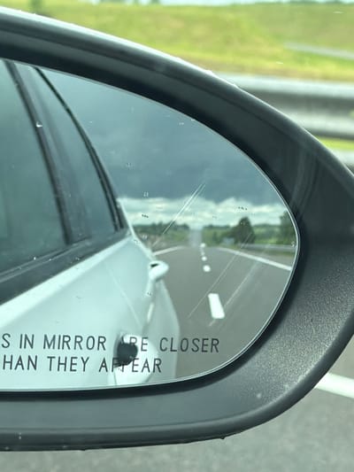 The image shows a close-up of a car's side-view mirror, reflecting a highway scene. The mirror displays text that reads "OBJECTS IN MIRROR ARE CLOSER THAN THEY APPEAR". The reflection shows a road with white lane markings, green fields, and trees on either side. The sky is cloudy. The mirror itself has some water droplets and fine scratches on its surface. The exterior of the car, a silver sedan, is visible on the left side of the image. The location is specified as Пяршайскі сельскі Савет, Belarus. There are no people visible in the image, and no actions or interactions are taking place. The environment appears to be rural, with a focus on the road and surrounding landscape as seen through the mirror.