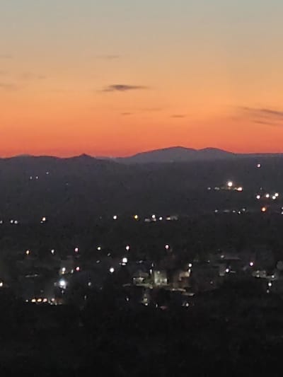 The image depicts a twilight view of a landscape, likely a city or town, with rolling hills and a dusky sky. The sky transitions from a pale blue at the top to a vibrant orange-red near the horizon, suggesting sunset or sunrise. Scattered clouds are visible in the upper portion of the sky. Below the sky, dark silhouettes of mountains or hills form the backdrop. In the foreground, lights from buildings and streetlights twinkle across the dimly lit landscape, indicating a settlement. The overall scene conveys a sense of peacefulness and transition from day to night. The specific location is not identifiable from the image alone, but the prompt suggests it is the city of Međugorje, Bosnia and Herzegovina.