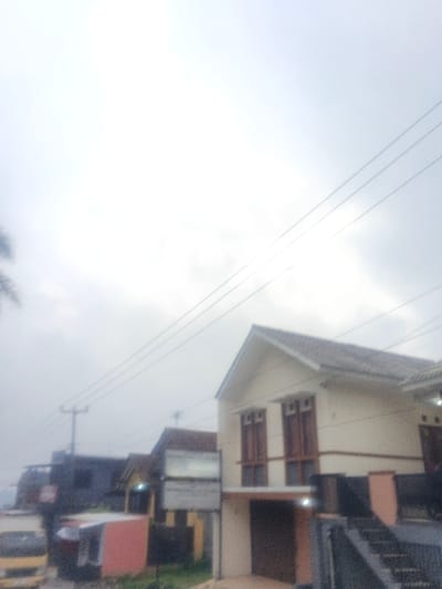 This image depicts a street scene in Dakshin Hichachhara, India, under an overcast and bright sky. The foreground on the right features a two-story residential or commercial building with a light beige or cream-colored facade, a prominent dark-tiled gable roof, and two tall, narrow wooden-framed windows on the upper floor. The ground floor displays a dark-colored entrance or garage door. To the immediate right of this building, a set of dark-colored stairs with a railing ascends upwards. Multiple horizontal utility lines traverse the upper portion of the frame against the bright sky. On the left side, a utility pole with several attached lines is visible. Beyond the pole and to the left of the prominent building, a series of other structures, varying in color (including dark grey, brown, and yellow), recede into the blurry background, suggesting a densely built area. At ground level on the far left, a portion of a yellow truck is visible. Adjacent to the truck, a small, reddish, single-story structure is present. A multi-paneled signpost stands in the middle ground, near the center-left. A partial palm frond is faintly discernible on the far left edge of the image. No individuals are visible, nor are any explicit actions or interactions. The overall scene appears hazy or slightly out of focus.