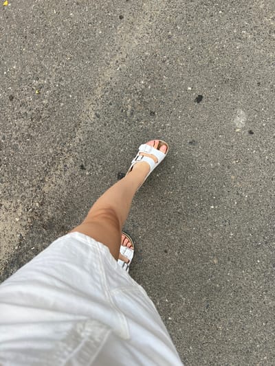 A person's legs and feet are shown from a top-down perspective, walking on a textured gray asphalt surface. The person is wearing a white dress that extends into the lower left corner of the frame, and white Birkenstock-style sandals on both feet. The asphalt has a rough, pebbly texture with scattered small debris. The scene is outdoors, likely in a city, but the specific location in Belozernyi, Russia, cannot be determined from the visual information alone. The lighting suggests it is daytime.
