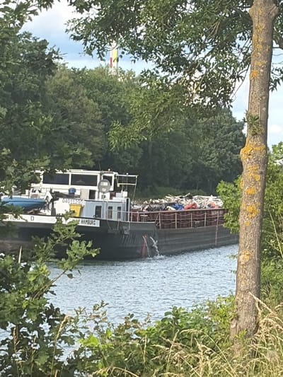 The image depicts a barge laden with debris being transported on a body of water. The barge is positioned in the foreground, with its cargo visible at the stern. The barge has the name "HAMBURG" printed on its side. The surrounding environment is lush with trees and vegetation, suggesting a natural or semi-natural setting. In the background, a tall structure with red, yellow, and white bands is partially visible above the trees, possibly a radio tower or similar landmark, which could help in pinpointing the exact location within Garbsen, Germany. The water appears calm, with gentle ripples indicating movement. The lighting suggests it is daytime, with sunlight filtering through the trees. There are no discernible people in the image.