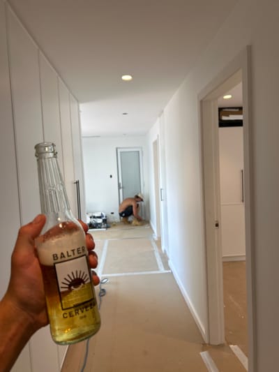 A person holds a half-full bottle of Balter Cerveza beer. An open doorway frames the background where a worker wearing a hard hat is kneeling on the floor. The interior space shows recently finished walls and flooring, with visible masking tape and construction debris. The perspective is an overhead view, giving a sense of depth to the hallway leading to a bright window. The color palette is dominated by whites, beiges, and the amber hue of the beer.