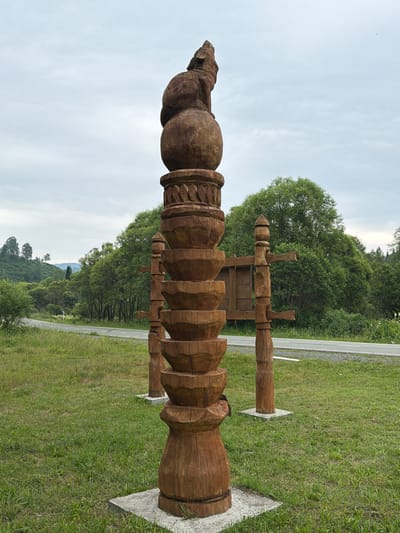 A wooden totem pole with a wolf-like animal carved at the top stands in a grassy field. The totem is made of stacked, bowl-shaped segments with decorative carvings at the top. To the right of the main totem are two smaller wooden structures. In the background, a road curves through a landscape of trees and rolling hills under a cloudy sky. The location is Srednie Kichi, Russia.