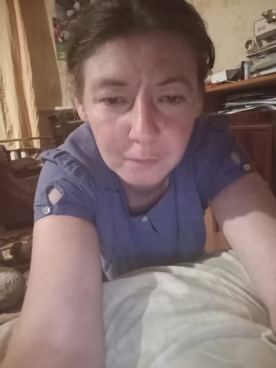 A medium shot, selfie-style image shows a woman with dark hair and fair skin indoors. She is wearing a light blue collared shirt with short sleeves that have cutouts and buttons. Her head is tilted down, and she is looking slightly off-camera with a neutral expression. Her arms are resting on what appears to be a light-colored duvet or bedspread. In the background, there is a curtain with vertical stripes, a wooden dresser or cabinet filled with papers and objects, and a wall with a picture. The lighting in the room is dim and warm. The setting is a domestic interior, likely a bedroom or living room, in Berezivka, Ukraine.