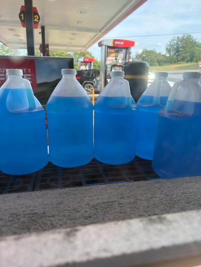The image shows a close-up of several plastic jugs filled with blue liquid, likely windshield washer fluid, lined up on a black plastic grate. The jugs have white caps and some have visible condensation on their sides. In the background, a gas station canopy with signs displaying the numbers "1" and "2" is visible, along with a red gas pump. A black SUV is parked in the background. The scene appears to be outdoors, under a cloudy sky, suggesting it is a gas station in Raphine, United States. The focus is on the jugs of fluid.