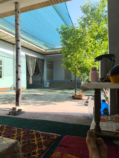 This image depicts an outdoor courtyard in Paytugh, Uzbekistan, bathed in bright sunlight. A large, blue mesh sunshade is stretched overhead, supported by a rustic wooden post in the foreground. To the right, a leafy green tree is visible against a clear blue sky. In the foreground, a person's feet are resting on a dark red cushion, partially covering a patterned rug. A low table to the right displays a pink thermos, a yellow object, a water bottle, and other various items. Sandals are placed on the ground near the wooden post. The background shows a building with light blue walls, a window, and drawn curtains. Further back, there is a pile of gravel and some bags. The ground is paved with concrete.