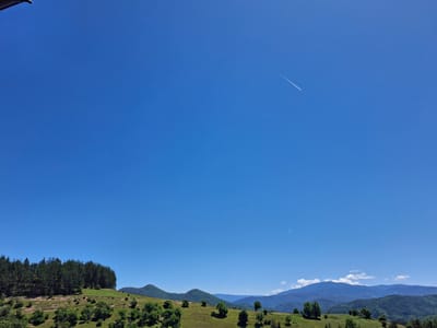 The image shows a landscape view in Alendarova, Bulgaria, under a clear blue sky. The terrain is composed of rolling hills covered in green grass with scattered trees. A dense forest patch sits on the left side of the frame atop one of the hills. In the distance, a mountain range is visible with some cloud cover near the peaks. Two contrails, likely from airplanes, are seen in the sky. There are no people visible in the scene. A portion of a building is visible in the top left of the frame.