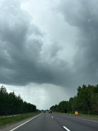 The image depicts a road in Голацкі сельскі Савет, Belarus, under a cloudy sky. The road is lined with trees on both sides. There are cars driving on the road. A yellow speed limit sign is visible on the right side of the road. The sky is predominantly grey and overcast, suggesting inclement weather.
