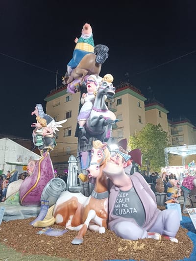 The image depicts a large, multi-tiered satirical monument, likely a Falla, brightly illuminated at night in El Port De Sagunt, Spain. The monument stands on a base of brown shredded material.
In the foreground, a stylized brown and white horse figure lies on its side, appearing surprised with an open mouth. Beside it, a male figure with exaggerated features, a blue and white checkered bandana, and a grey t-shirt reading "KEEP CALM AND THE CUBATA" sits, grinning upwards. A silver barrel and a silver fire hydrant-like object are positioned near these figures. To the left, a figure draped in purple fabric is partially visible.
Above the foreground figures, on a higher platform, a large, exaggerated female figure with white hair and glasses rides a grey horse, both appearing agitated. To her left, another platform supports an angelic figure with white wings carrying a green-striped object, beside a figure with a prominent nose and dark hair.
At the apex of the monument, a stout male figure in a light blue shirt, green and yellow sash, and blue pants is precariously balanced or falling from a brown horse figure.
In the background, a multi-story building with light brown facades and green trim on its rooflines is visible under a dark sky. Numerous indistinct figures of people are present on both sides of the monument, suggesting a public gathering. A large white tent structure is on the far left, and part of a metal truss structure with a tree is on the right. The scene is lit by artificial lights. Several informational signs are visible at the base of the monument.