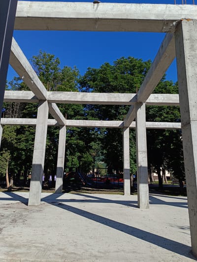 The image shows the skeletal framework of a concrete building under construction. Four large concrete pillars support a grid of horizontal concrete beams, forming a cubic structure. The construction site appears to be an outdoor area with a clear blue sky overhead. Behind the concrete structure, a dense line of green trees forms a backdrop, suggesting a park or residential area. In the distance, through the gaps in the framework, a children's playground with a slide is visible, along with some park benches. The ground in the foreground is made of concrete and is partially covered with loose gravel and debris. Sunlight casts long shadows from the concrete pillars and beams across the ground, indicating it is daytime. There are no people clearly visible in the frame.