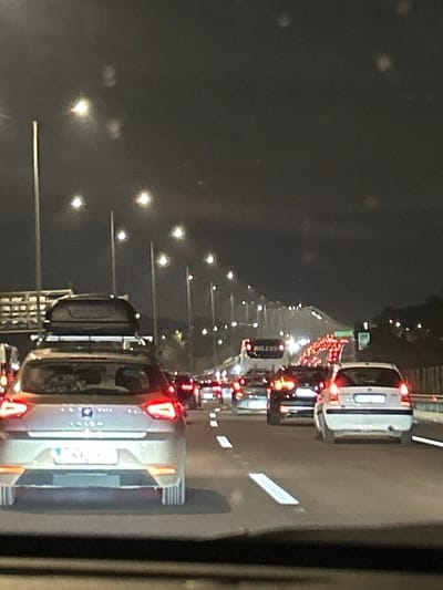 The image depicts a nighttime scene of a congested multi-lane highway or major road, viewed from a slightly elevated perspective. The road stretches into the distance, filled with numerous vehicles forming a long line of traffic, suggesting a jam or very slow movement.

The primary subjects are the cars and trucks, with their bright red taillights creating a prominent visual feature that recedes into the background. To the left of the road, a consistent series of streetlights illuminates the scene, their bright points of light also extending into the distance and casting some reflections and lens flares across the dark sky. White lane markings are visible on the asphalt.

In the foreground, the rear of a light-colored (possibly silver or grey) SUV is visible, equipped with a roof rack holding luggage. Its taillights are prominently lit. Further down the road, a white car and a dark commercial vehicle, possibly a truck or van, are discernible. The word "BLESS" is visible in white text on the rear of the dark commercial vehicle in the mid-ground.

The activity taking place is night-time driving in heavy traffic. The time of day is clearly night, indicated by the darkness and reliance on artificial lighting from vehicles and streetlights. The location context is Anatoliki Attiki, Greece. No individuals are explicitly visible, but their presence in the vehicles is implied.
