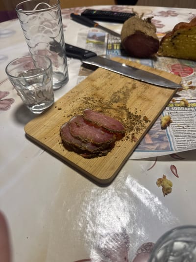 An overhead shot reveals a table setting in Maslarevo, Bulgaria. A white patterned vinyl tablecloth with pinkish-purple floral motifs covers the surface. On the table, a light-colored rectangular wooden cutting board is positioned, bearing multiple thin slices of dark-crusted, pinkish-red cured meat. Dark granular spices are scattered on the board around the meat. A silver-bladed knife with a black handle rests atop the cutting board. Adjacent to the board, a tall, clear glass with a wavy texture and a smaller, clear glass containing a translucent liquid are present. Behind the cutting board, a whole, dark-brown, irregularly shaped piece of cured meat, possibly a dried sausage or loin, wrapped in a fibrous casing, sits beside a partial section of yellow bread or cake containing dark inclusions. Crumbled pieces of yellow bread or cake are scattered across the tablecloth, particularly to the right of the cutting board. In the upper background, a dark remote control is visible on the tablecloth. The scene depicts a domestic setting with prepared food items.