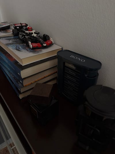 This is a close-up, angled shot of a bookshelf in what appears to be a home setting. The subjects are primarily objects. In the foreground, several books are stacked, some with colorful illustrated covers, and others with blue spines bearing Cyrillic text. A black and red toy race car, possibly a model of a Formula 1 car, is perched on top of one of the books. The car features branding that includes "JJAS," "HAAS," "Money," and "Gram."

Further back on the shelf, a dark blue desk organizer with labels for each day of the week (Monday through Sunday) is visible. The word "BONO" is printed prominently on the organizer. To the right of the organizer, a dark object, possibly another book or a decorative item, is situated. The overall scene suggests a personal space, perhaps a study or living room. The lighting appears to be artificial, indicating it is likely indoors. There are no people or animals visible in the image.