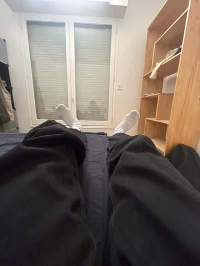 The image depicts an interior scene, likely a room in Boulogne-Billancourt, France, from a first-person perspective looking down. The lower half of an individual's body is visible, dressed in dark trousers or sweatpants and white socks, lying supine on a bed covered with a dark blue sheet. In the midground, a large, white-framed double window with closed horizontal blinds or shutters occupies the central wall. A faint, dark silhouette, possibly of a person, is discernible behind the blinds. To the left of the window, a portion of a light-colored vertical storage unit or wardrobe is visible, with clothing draped on it. To the right, a light-colored wooden shelving unit with multiple compartments holds various items, including a piece of light-colored fabric on an upper shelf. A light-colored electrical outlet or switch is mounted on the wall between the window and the shelving unit. The walls are light-colored, and the overall lighting is interior ambient.