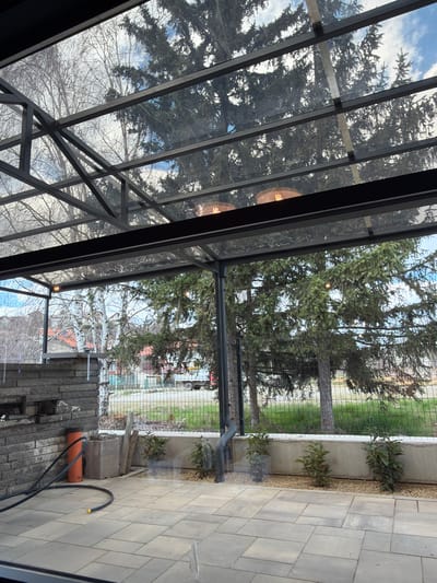 The image depicts an outdoor patio or enclosed area in Rudartsi, Bulgaria, likely part of a building. The patio features a glass and metal frame structure overhead, allowing a view of trees and sky. The flooring consists of grey paving stones. Along the side, there is a low concrete wall with plants. A black hose and an orange pipe are visible on the ground. A metal fence can be seen beyond the concrete wall, with trees and buildings visible in the background.