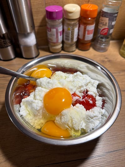 The image shows a stainless steel bowl filled with ingredients for a meal, likely a breakfast or a baking preparation. The bowl contains several whole eggs with their yolks intact, separated from the whites. There are also dollops of a white, cottage cheese-like substance and some chopped red ingredients, possibly tomatoes or preserved fruit. A silver spoon is partially submerged in the mixture, suggesting it is being stirred or prepared. In the background, on a wooden surface, are several spice grinders. One appears to be filled with salt, and another with black peppercorns. Other spice containers are visible, one with a pink cap, another with a beige cap, and one with an orange cap, along with a glass jar labeled "Pfeffernuss". The scene is indoors, with a wooden countertop visible. This image was captured in Kirtorf, Germany.