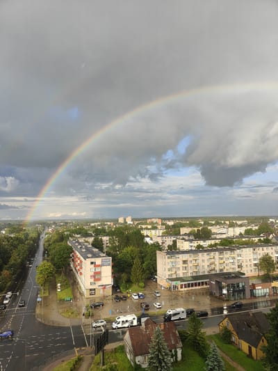 The image captures an aerial view of a city scene in Panevėžys, Lithuania, dominated by a prominent rainbow stretching across a cloudy sky. The foreground features a street intersection with wet asphalt, indicating recent rainfall, and several vehicles, including a white camper van, cars, and a blue sedan. The mid-ground displays apartment buildings and smaller houses, some with red tiled roofs. A multi-story apartment building with balconies is visible on the left side of the intersection. Trees line the streets and surround some of the buildings. In the distance, more apartment complexes and a treeline form the urban and suburban landscape. The sky is characterized by a mix of dark, heavy clouds and patches of lighter sky, with the rainbow appearing as a vibrant arc against this backdrop. A few people are visible on the sidewalks and near the buildings, engaged in pedestrian activities.