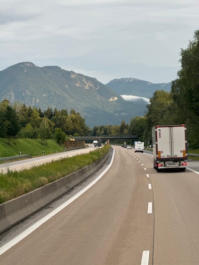The image shows a highway in Raubling, Germany. The highway is paved with asphalt and has white lane markings. There are several vehicles on the highway, including a large white semi-truck with the word "SCHMITZ" visible on the back. Other vehicles, including cars and smaller trucks, are also present. The highway is surrounded by lush green trees and a steep, mountainous landscape. The sky is overcast, suggesting a cloudy or foggy day. The scene appears to be a daytime shot, given the lighting. The overall impression is of a scenic drive through a mountainous region on a highway.