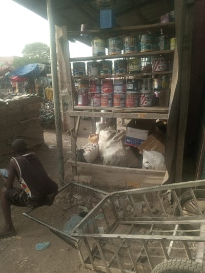 This image depicts an outdoor market stall in Kofar Ruwa, Nigeria. The scene is illuminated by daylight, with a bright sky visible in the upper left.
A central wooden-framed stall features multiple shelves stocked with numerous cans of paint in various colors and brands, including "Foco," "Citizen," "Sharom," "Makollex," "Boss," and "Glensilk." Below the shelves, on the dirt ground, are several large fabric sacks, multiple white plastic jerrycans, one yellow jerrycan, and cardboard boxes. One cardboard box shows blue and white markings; another features a black-bordered hazard diamond symbol. A blue container is suspended from the stall's roof structure.
To the left, a dark-skinned male individual is positioned with his back to the camera, appearing to squat or sit on a low, indistinct object. He wears a dark sleeveless top with a patterned lower back section and dark, possibly patterned, shorts. He holds a small, light-colored object in his hands, seemingly engaged with it.
In the lower-center and right foreground, an empty, silver-colored metal-framed handcart or pushcart rests on its side, revealing its internal structure and one wheel on the right. The ground throughout the foreground and midground is dirt, scattered with debris including small rocks and light blue plastic elements. In the background, to the left, informal structures are visible, including a structure partially covered by a bright blue tarp, and distant trees under the bright sky.