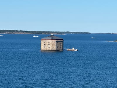 The image depicts a cylindrical, stone fort situated in the blue waters of the Baltic Sea, in the archipelago of Karlskrona, Sweden. The fort, which appears to be a historical defensive structure, has a dark, conical roof and several small, arched windows visible on its exterior. A small, connected platform with a white structure and what appears to be a small boat is attached to one side of the fort. The water is calm with gentle ripples. In the background, a long, tree-covered island coastline stretches across the horizon under a clear blue sky. Several small boats and a larger ferry-like vessel are visible in the distance, navigating the sea. The overall scene is peaceful and evokes a sense of historical significance and natural beauty.