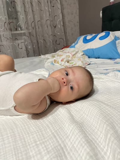 A newborn infant lies on a white, textured bedsheet, with its right hand in its mouth. The infant is wearing a white onesie. Behind the infant, a blue and white pillow with a large "S" logo is visible, along with a patterned blanket. The background includes a curtain with a swirling pattern and a dark headboard. The scene appears to be indoors, likely in a bedroom. The presence of a pillow and blanket suggests a domestic setting. There is no information in the image to determine its location within Fălești, Moldova.