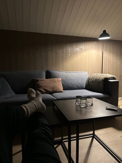 A cozy, slightly elevated view reveals a sofa against a light wood-paneled wall, illuminated by a black floor lamp. A patterned cushion rests on the sofa, beside a brown pillow. A dark-topped table, with three glasses, sits to the right. A person's legs, clad in dark pants and ribbed socks, are stretched out, suggesting relaxation. The scene is warmly lit, creating a comfortable and inviting atmosphere.