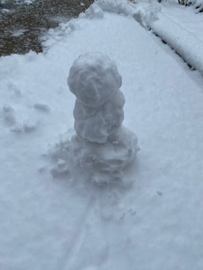 The scene, located in Yanaryk, Uzbekistan, depicts an outdoor environment under overcast lighting, extensively covered by a fresh layer of white snow. Centrally positioned, a small, crudely constructed snowman stands, composed of three irregularly shaped snowballs stacked vertically. The topmost snowball is the smallest, forming the head. The middle snowball is slightly larger, and the bottom snowball, serving as the base, is the largest and most amorphous, with loose snow collected around its foundation. The ground surrounding the snowman is uniformly covered in undisturbed snow. In the upper-left quadrant, a dark, textured surface, consistent with pavement such as asphalt or concrete, is partially visible where the snow cover is thinner. Along the right side of the image, a faint, dark linear edge, resembling a curb or a raised border, is partially obscured by snow. No individuals or animals are present, and no actions or interactions are depicted within the frame.