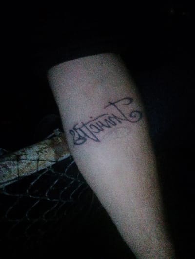 The image shows a tattoo on a person's arm, with a dark background. The tattoo is a word written in a cursive script, potentially in an Indian language, though specific identification is difficult due to image quality. The surrounding environment is dark and unclear, with what appears to be a fence partially visible. The location in Tahlequah, United States of America is not known from this image.